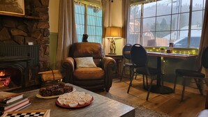 A cozy living room with a stone fireplace, a leather armchair, a dining table with chairs, and a view of the outside through large windows. - Cinnamon Bear Inn (Mammoth Lakes)