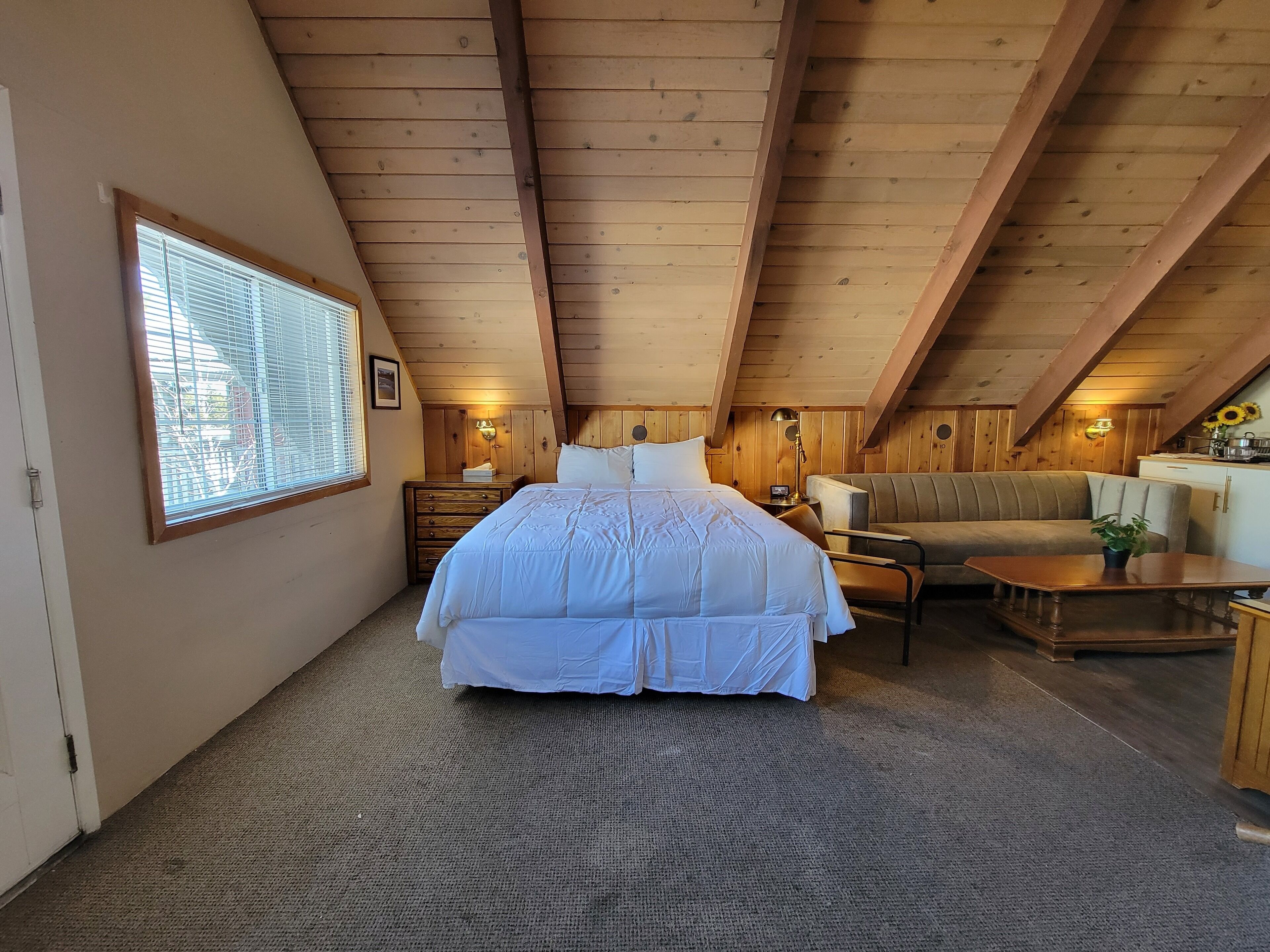 A cozy room with a large bed, a sofa, a small table, and a window with blinds.