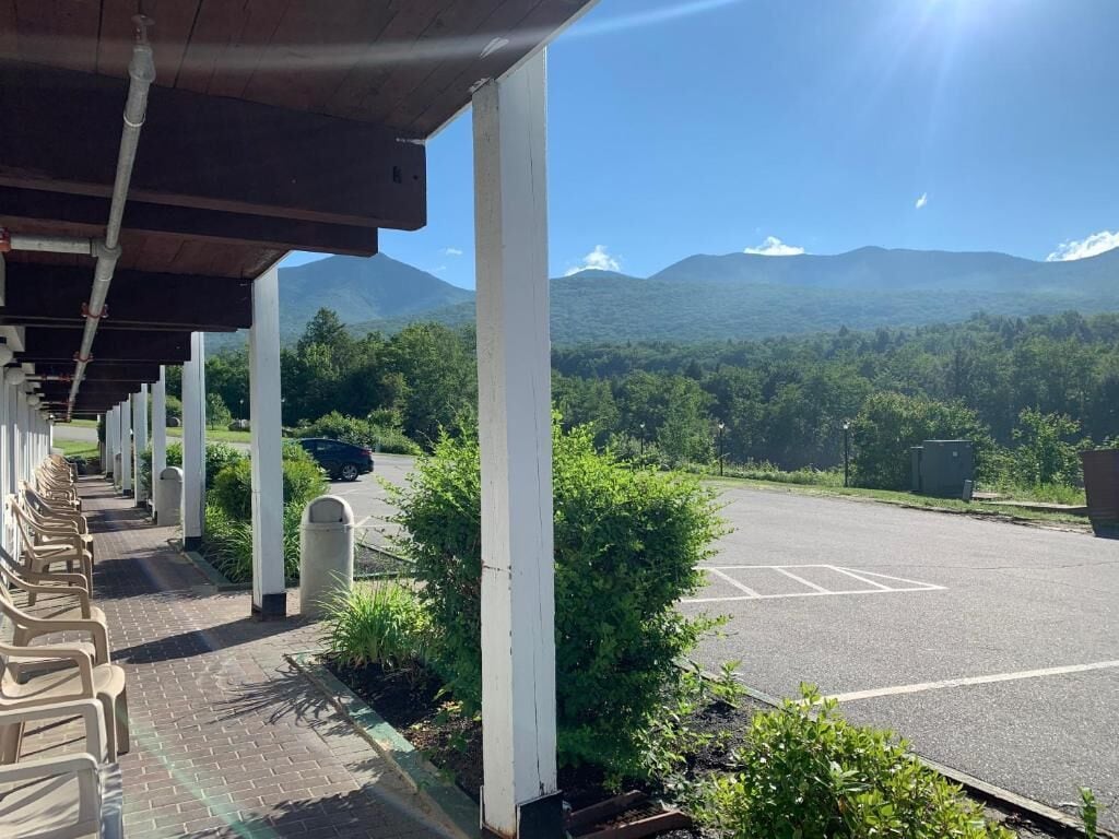 A covered outdoor area with a row of chairs, a parking lot, and a view of a forested mountain range.