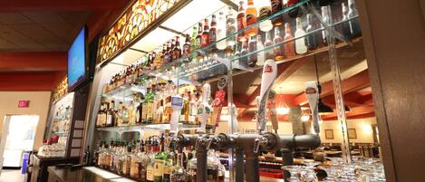 A bar area with multiple shelves of bottles and glasses, a television mounted on the wall, and a counter with a tap.