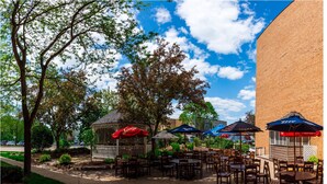 An outdoor patio area with tables and chairs, umbrellas, and a brick building in the background. - Hotel Mead Resorts & Conventions Center (Wisconsin Rapids)