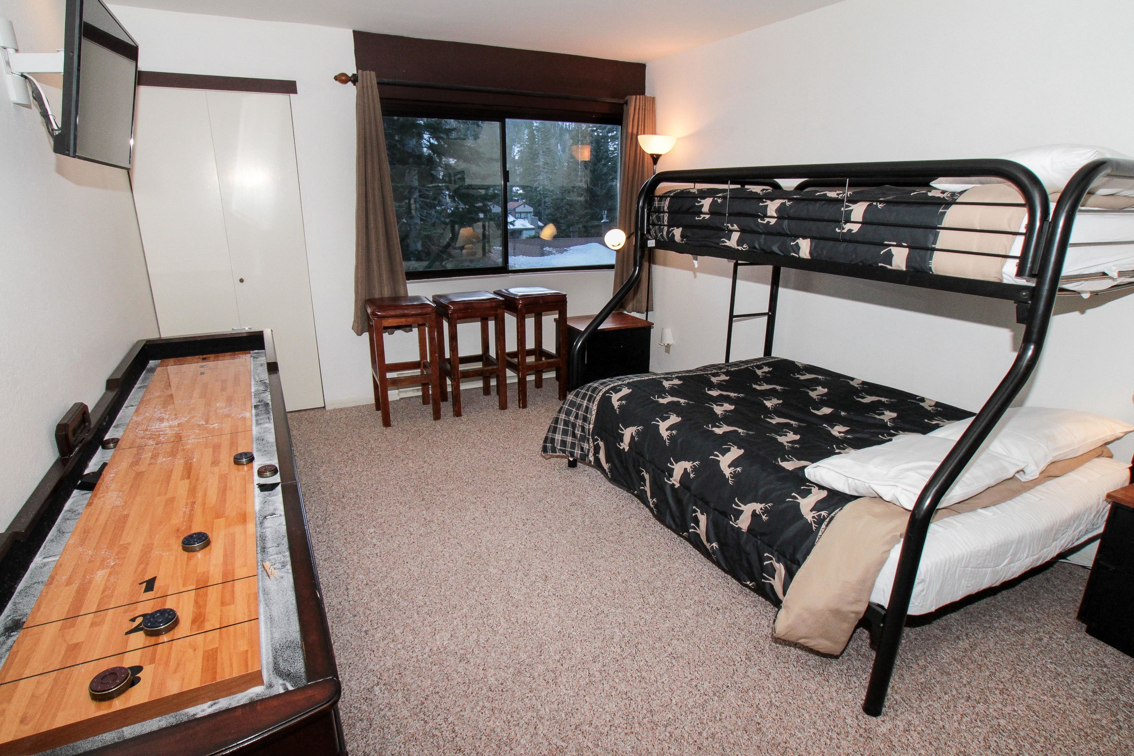 Ein Stockbett mit gemustertem Bettbezug, ein Holztisch mit Billardspieltisch und ein Fenster mit Blick auf Bäume.