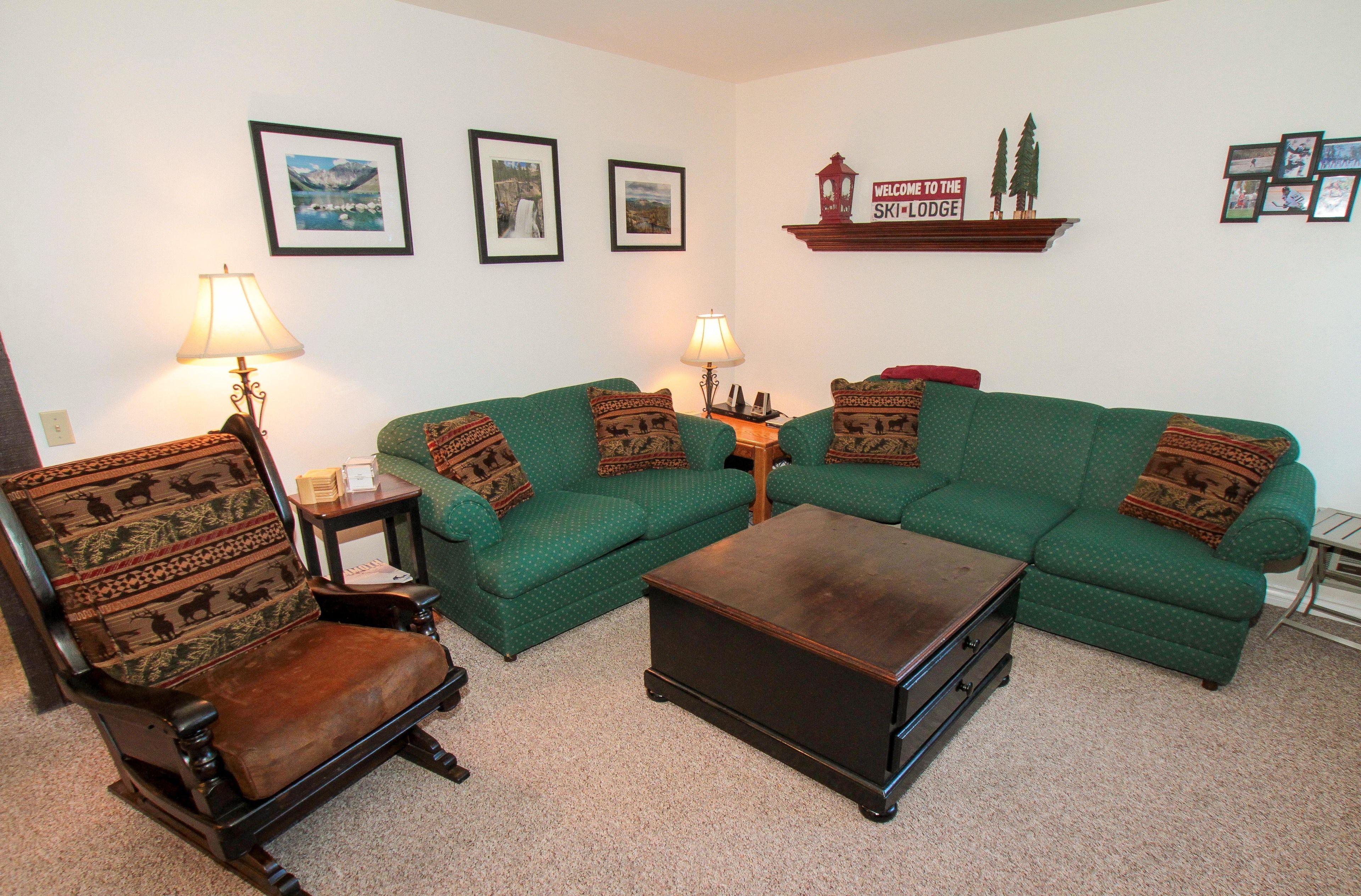 A living room with a green sofa set, a wooden coffee table, a wooden armchair, and two wall-mounted lamps.