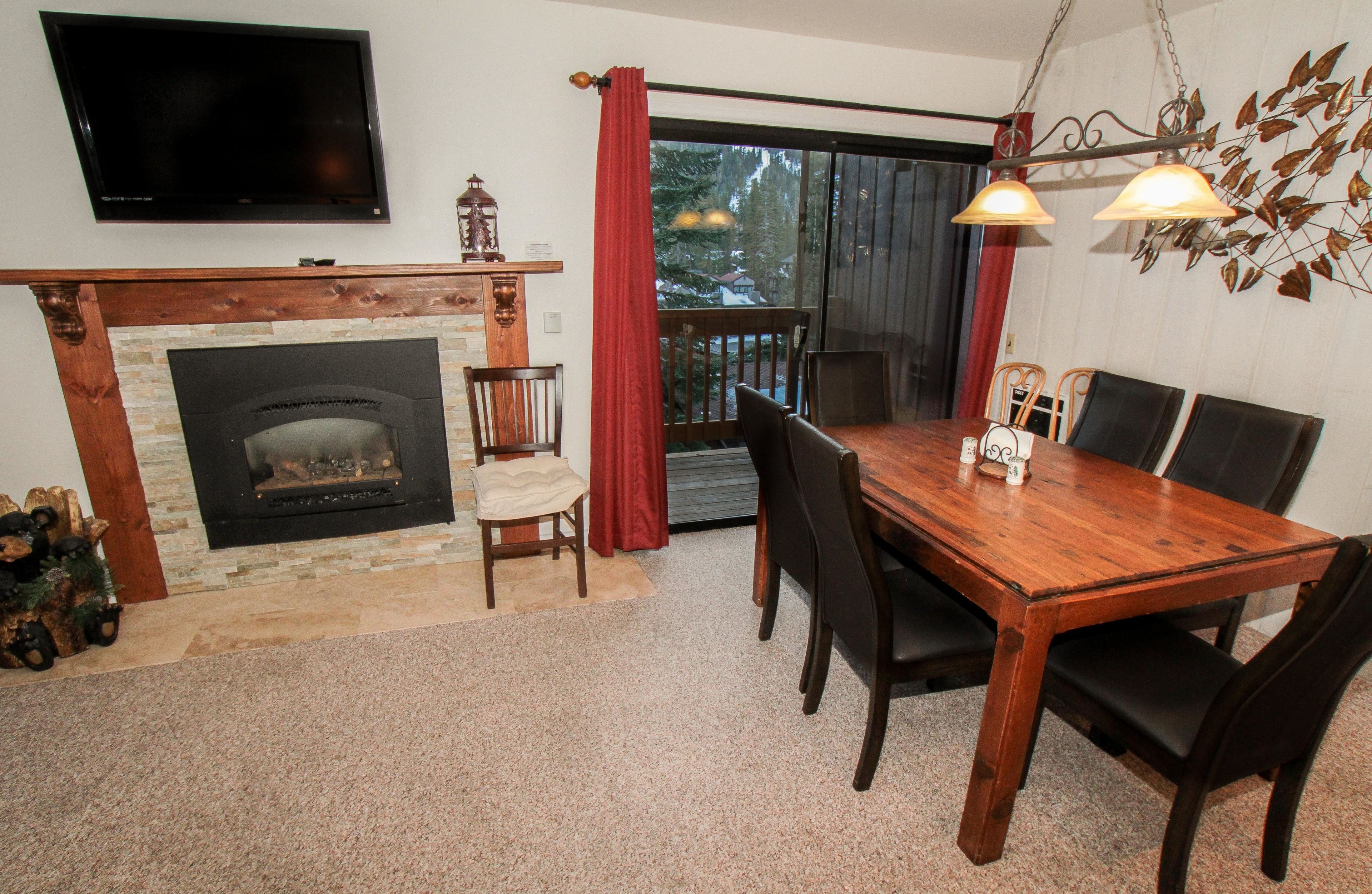 A dining area with a wooden table and chairs, a fireplace, a television mounted on the wall, and a view through a glass door.