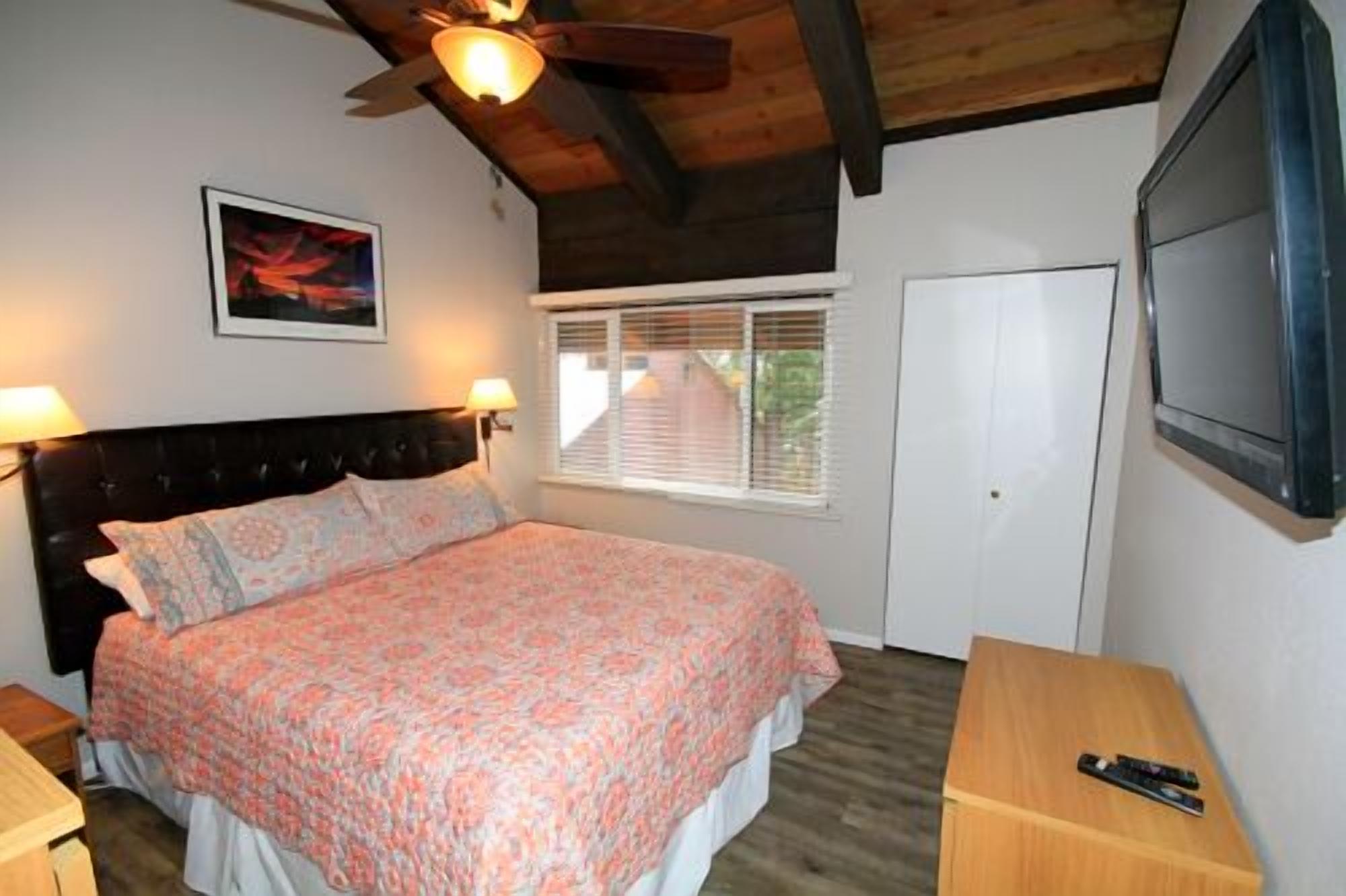 A bedroom with a bed, bedside lamps, a ceiling fan, a window with blinds, and a TV mounted on the wall.