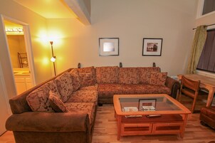 A living room with a sectional sofa, a glass coffee table, and a lamp. There are framed pictures on the wall and a window with curtains.