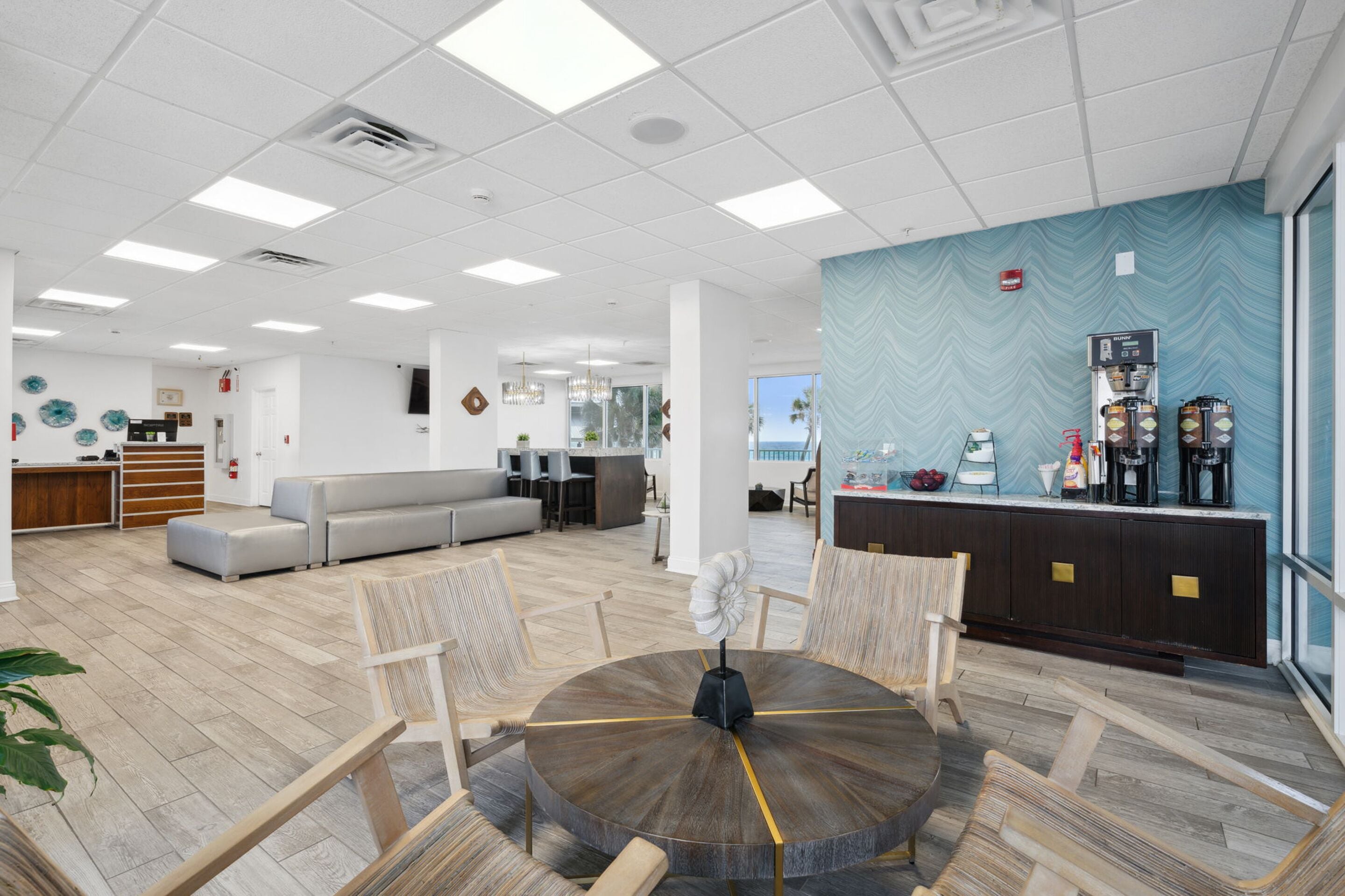 A modern lobby with a round wooden table, cushioned seating, a reception desk, and a view of the outdoors.