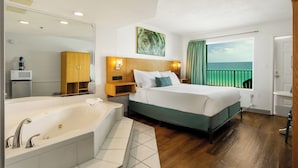 Quarto de hotel com uma cama grande, banheiro separado com banheira, um armário de madeira, um quadro e vista para o oceano.