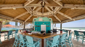 Uma área de bar ao ar livre coberta, com cadeiras turquesa, balcão de madeira e vista para a praia.