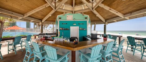 A covered outdoor bar area with turquoise chairs, a wooden bar counter, and a view of the beach.