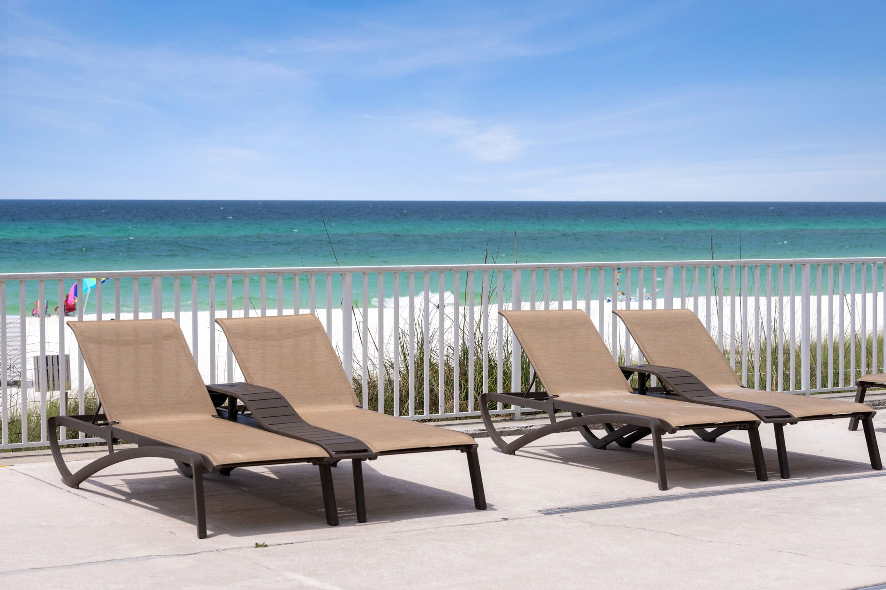 Vier Sonnenliegen mit Metallgestell und beiger Liegefläche stehen auf einer Betonplattform. Im Hintergrund ist ein weißer Zaun und der Blick auf das Meer zu sehen.