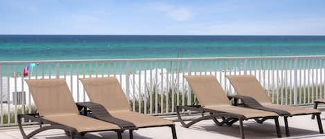 A row of four sun loungers with metal frames and beige fabric, positioned on a concrete deck with a view of the ocean and a white fence in the background.