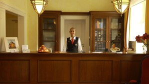 Un mostrador de recepción con una recepcionista, paneles de madera, cuadros enmarcados y un jarrón con flores.