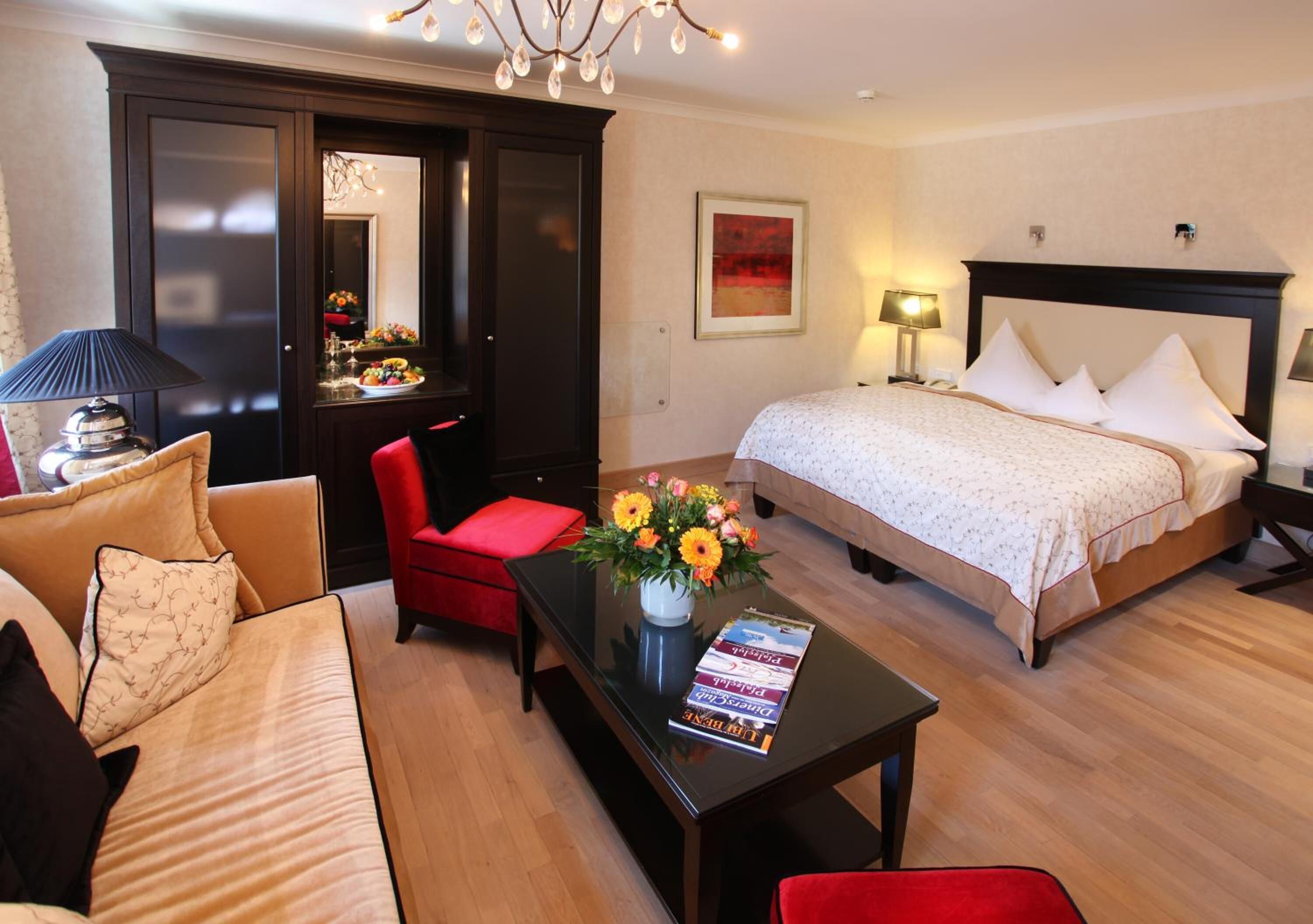Una habitación de hotel con cama, sofá, una silla roja, una mesa de centro con un florero y un escritorio con lámpara.