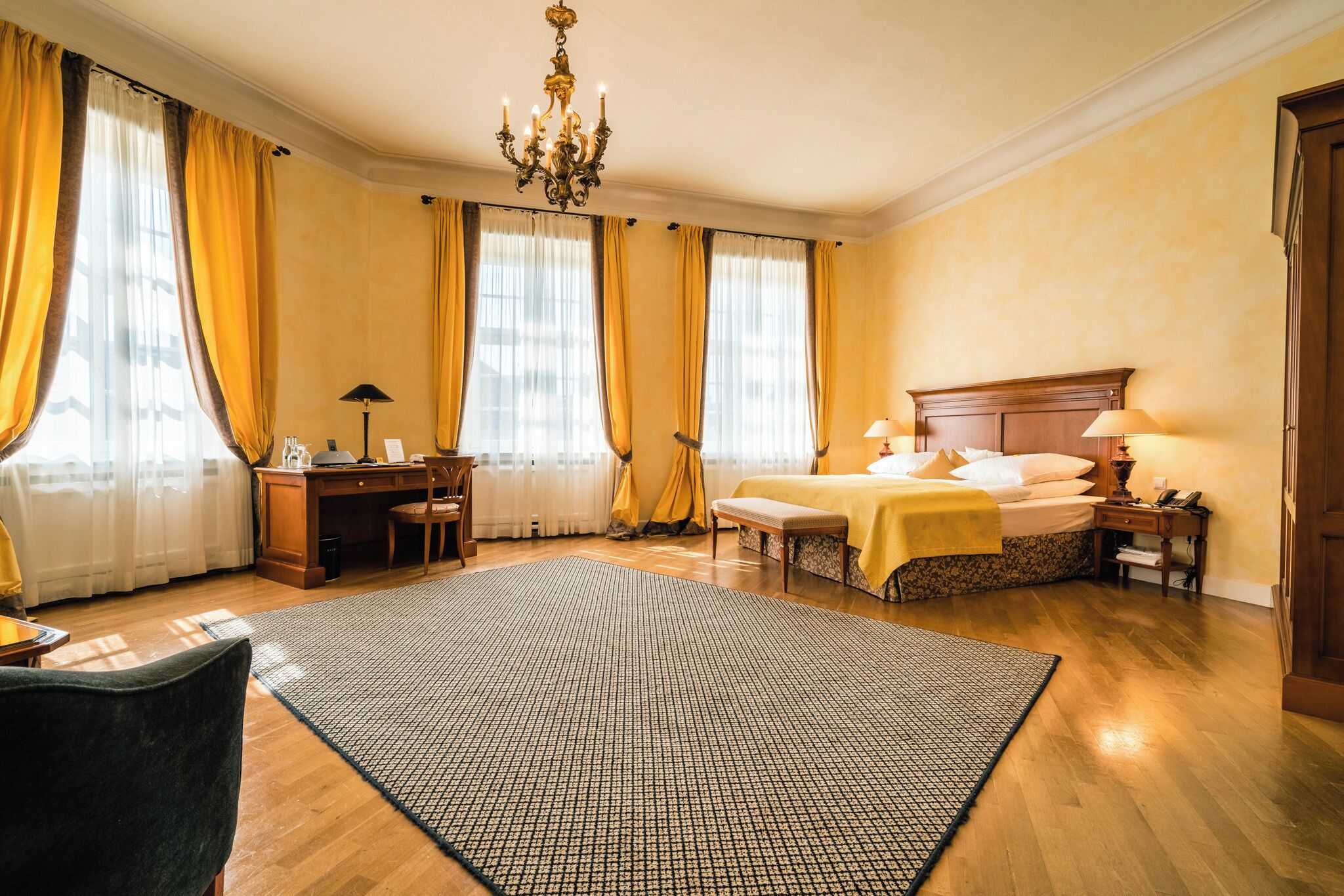 A hotel room with a wooden floor, a large bed with a headboard, a desk with a chair, a desk lamp, and a chandelier.