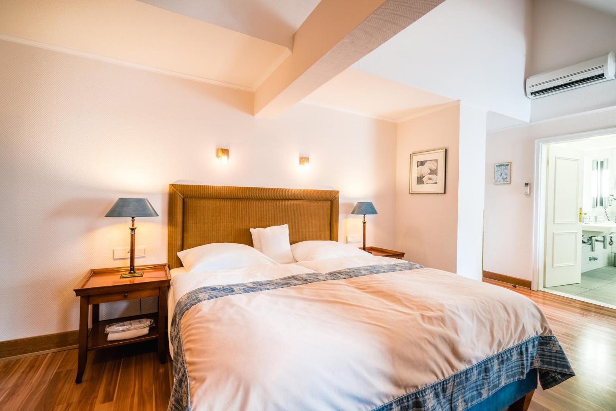 Quarto de hotel com uma cama grande, duas luminárias de cabeceira, uma cabeceira de madeira e um aparelho de ar condicionado.