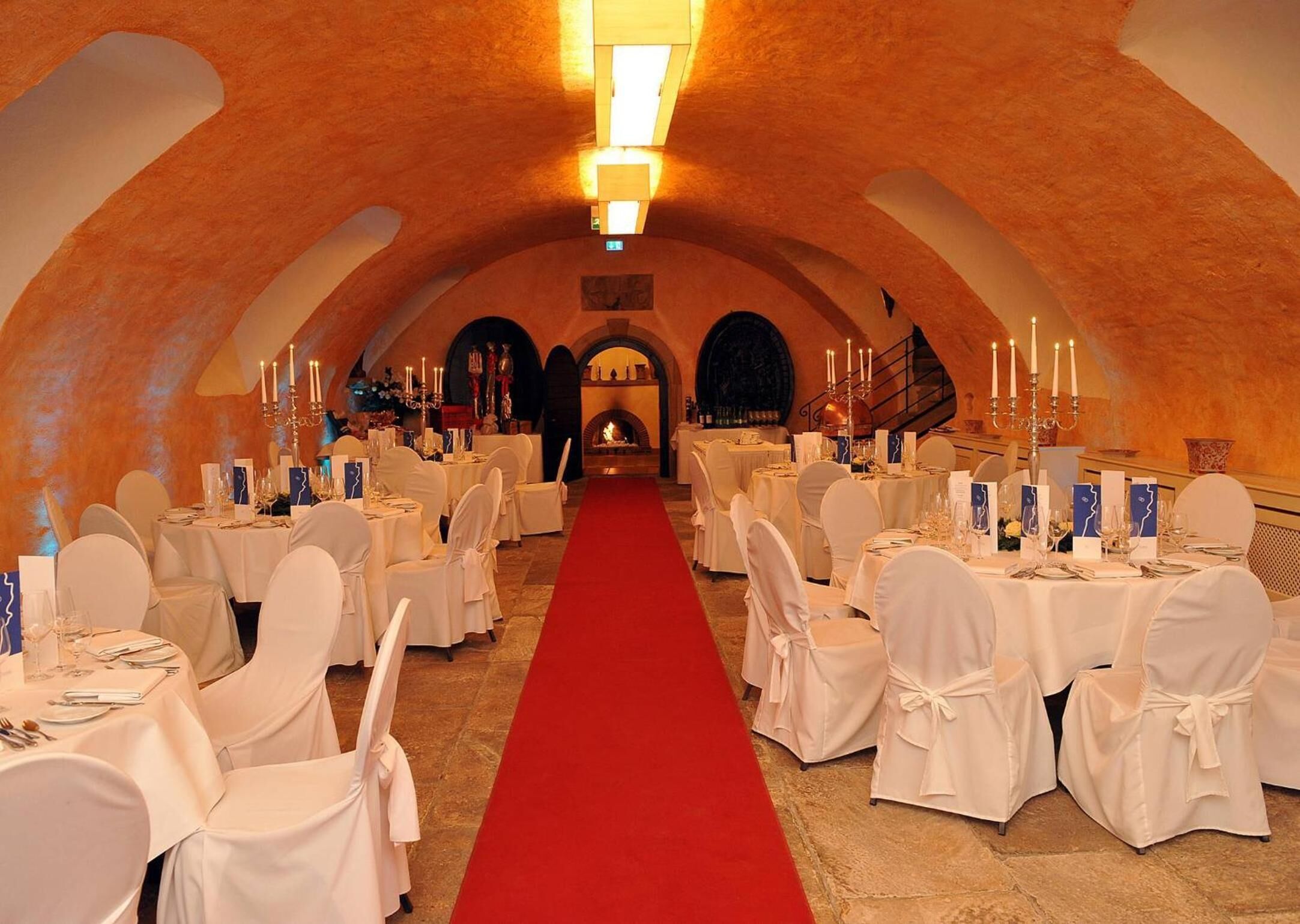 A banquet hall with white tablecloths, round tables, and chairs, a red carpet, and a high arched ceiling.