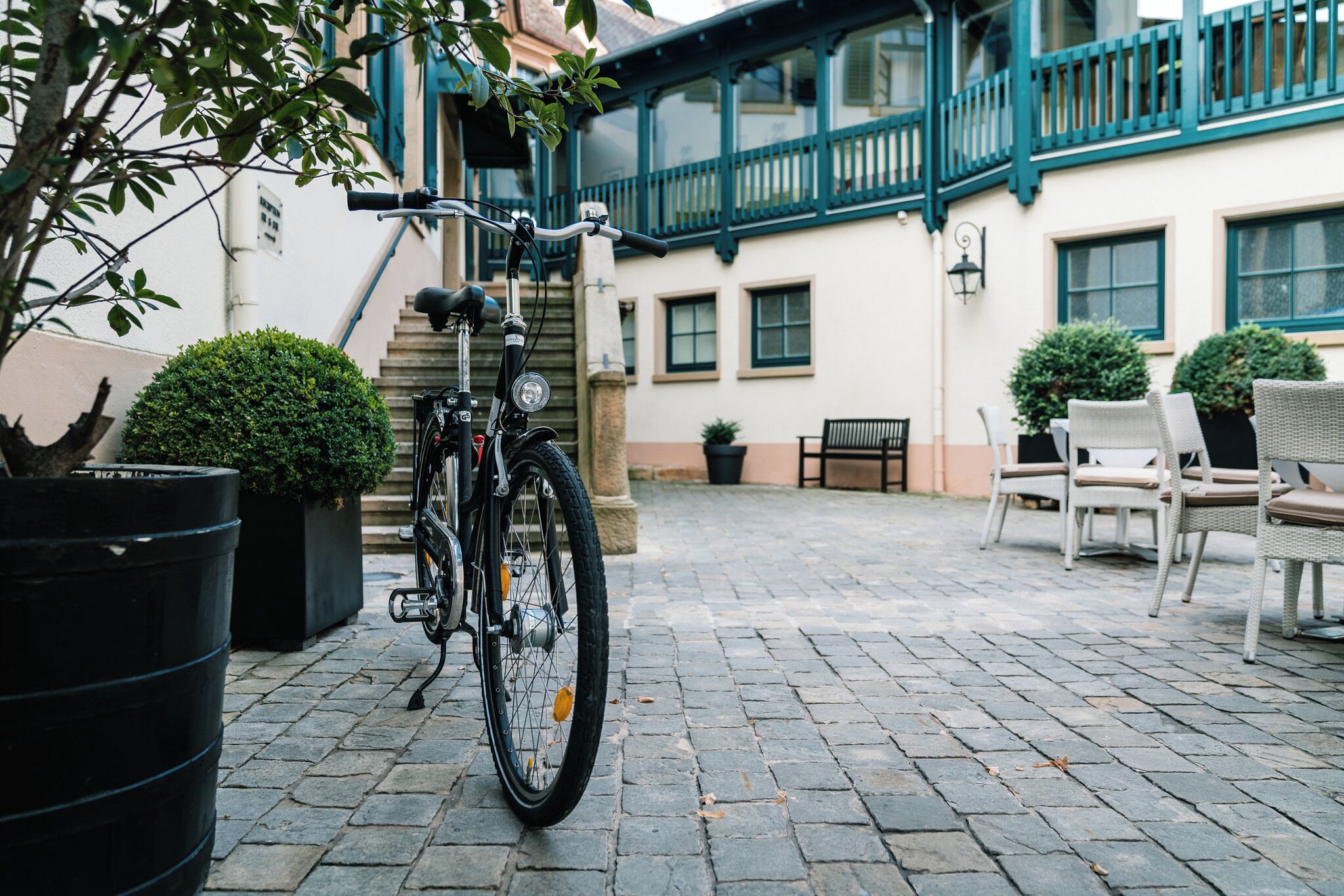 A bicycle parked in front of a building with a balcony, potted plants, and outdoor seating.