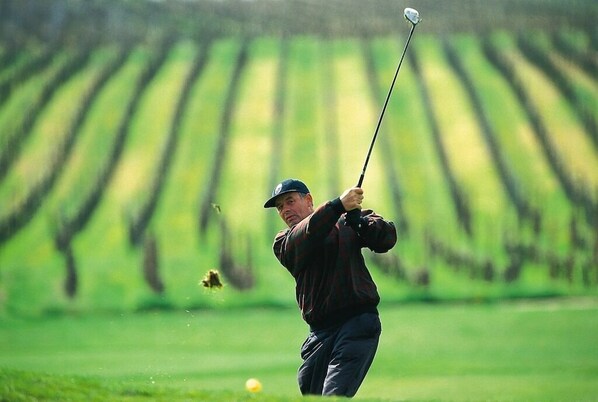 Un golfista en movimiento sobre un campo de golf, con filas de cultivos al fondo.