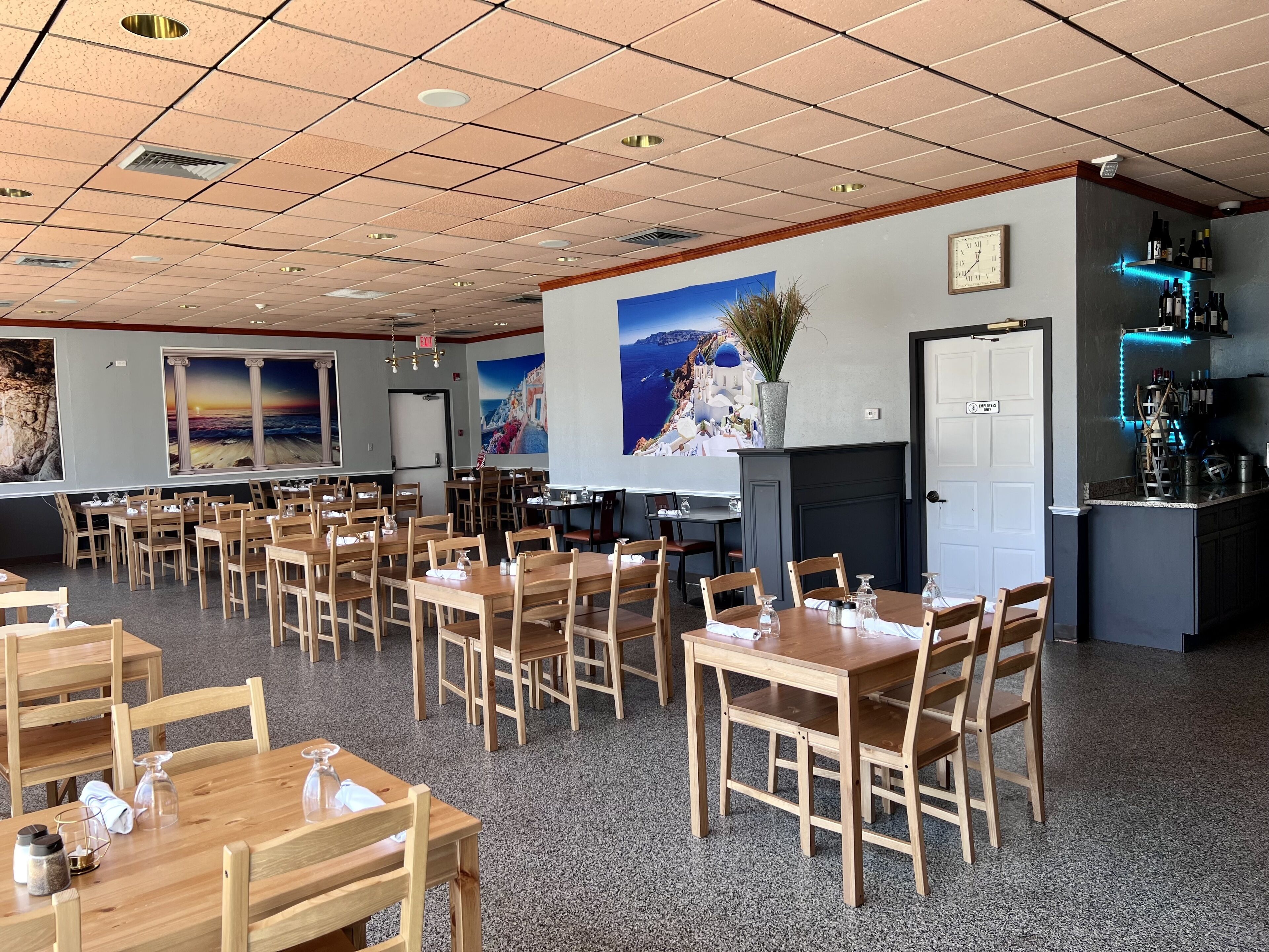 A restaurant with multiple wooden tables and chairs, a kitchen area with wine bottles, and a wall with a sunset painting.