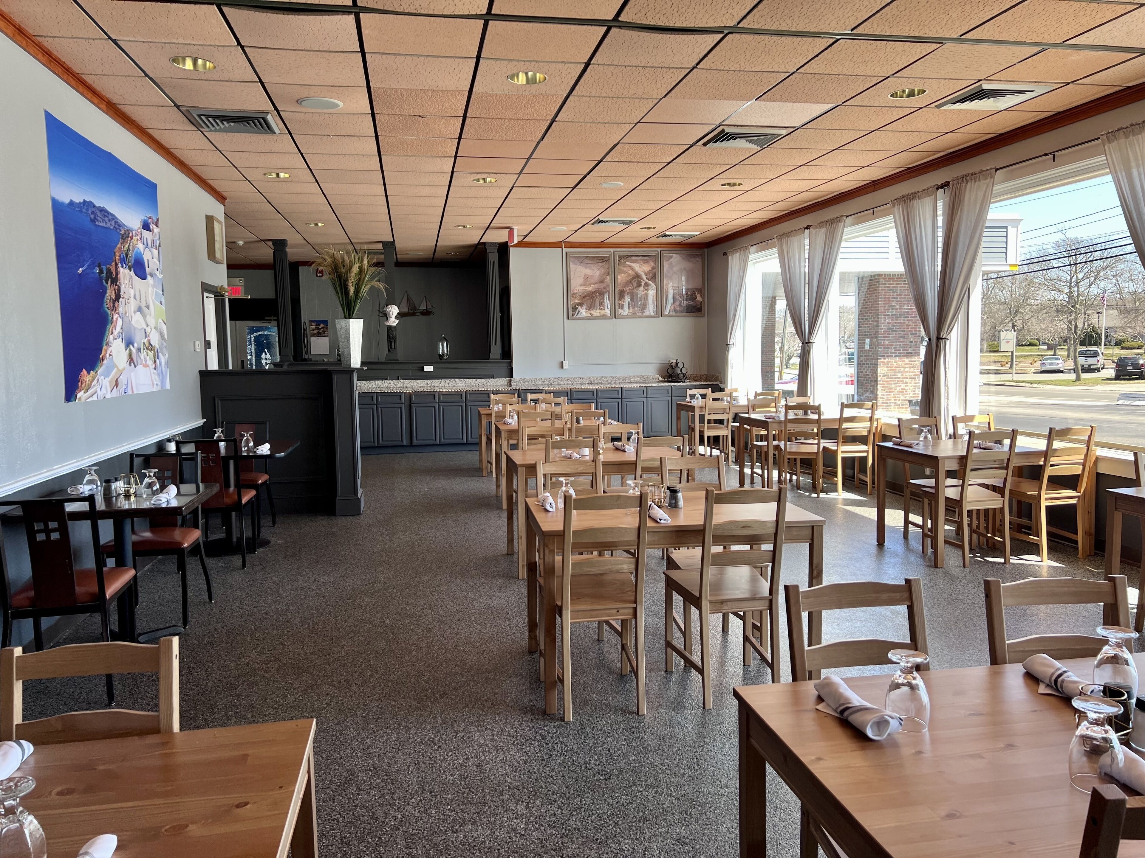 A restaurant with wooden tables and chairs, a kitchen area, and large windows with curtains.