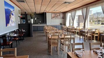 A restaurant with wooden tables and chairs, a kitchen area, and large windows with curtains.
