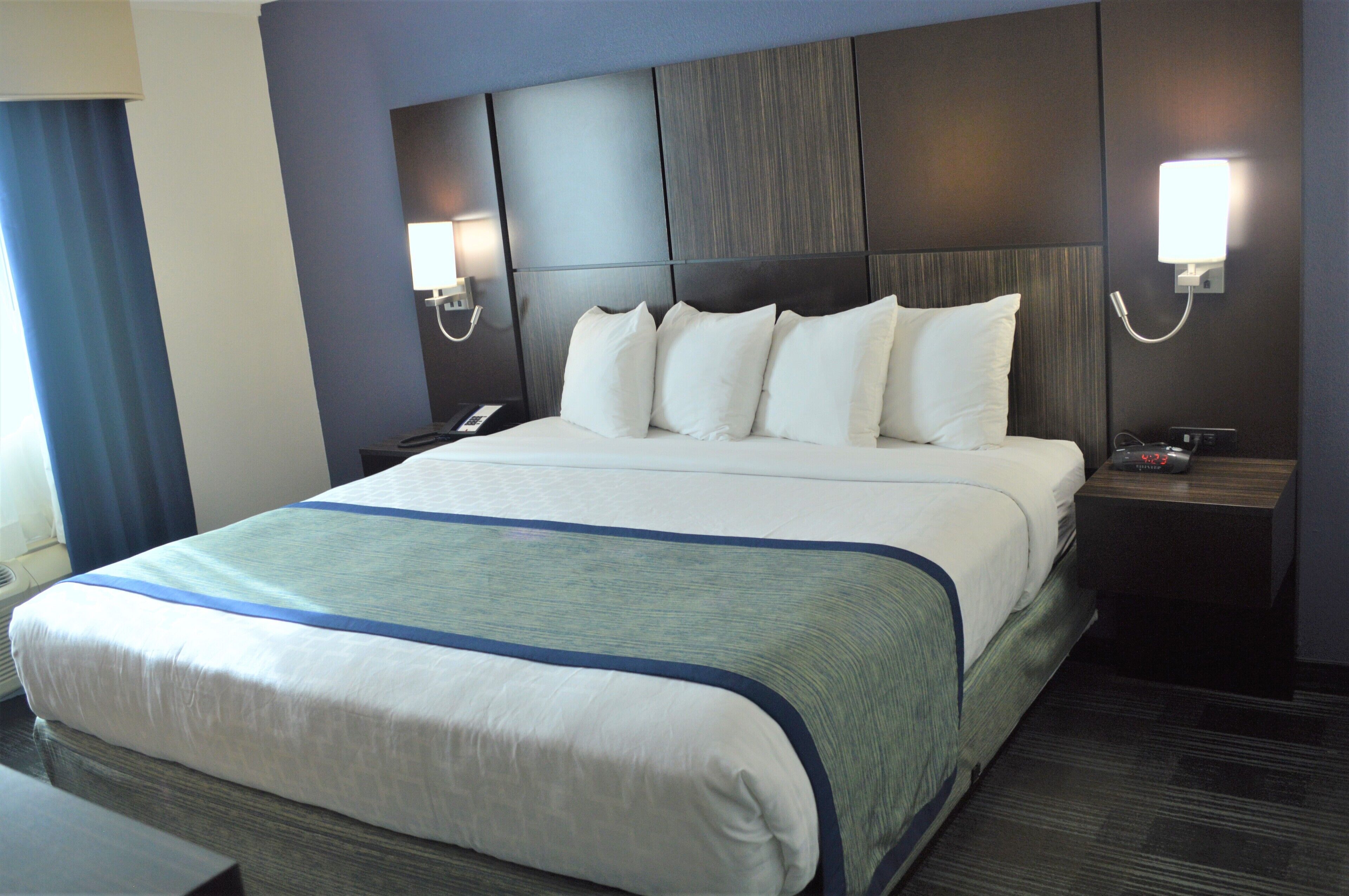 A hotel room with a large bed, a nightstand with a phone and a clock, and two wall-mounted lamps.