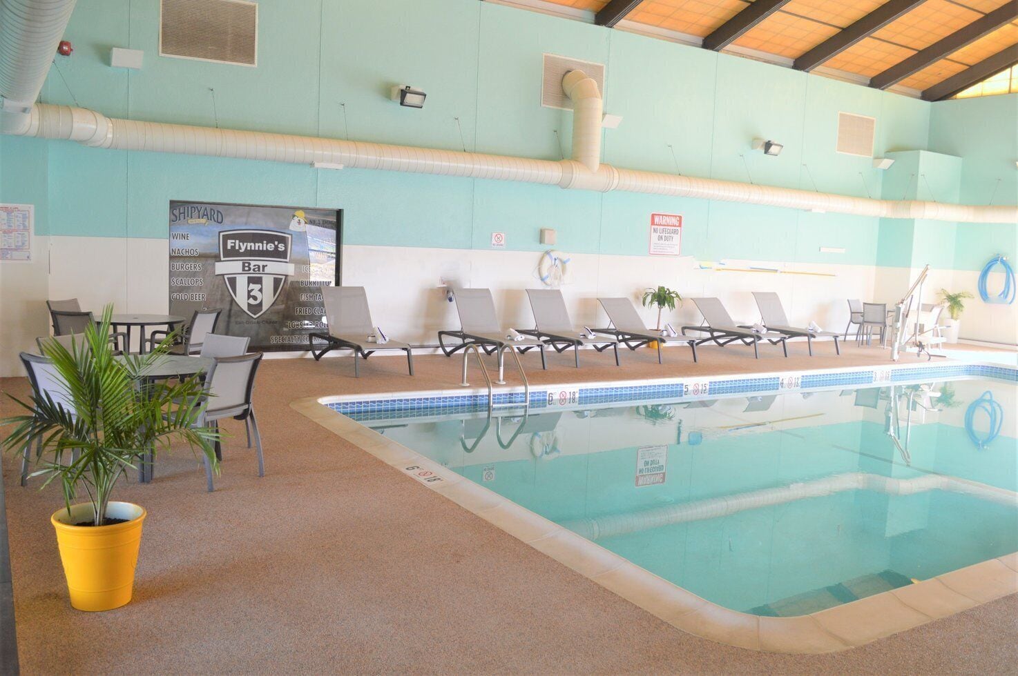 An indoor swimming pool with lounge chairs, a table with chairs, and a menu board advertising Flynie's Bar.
