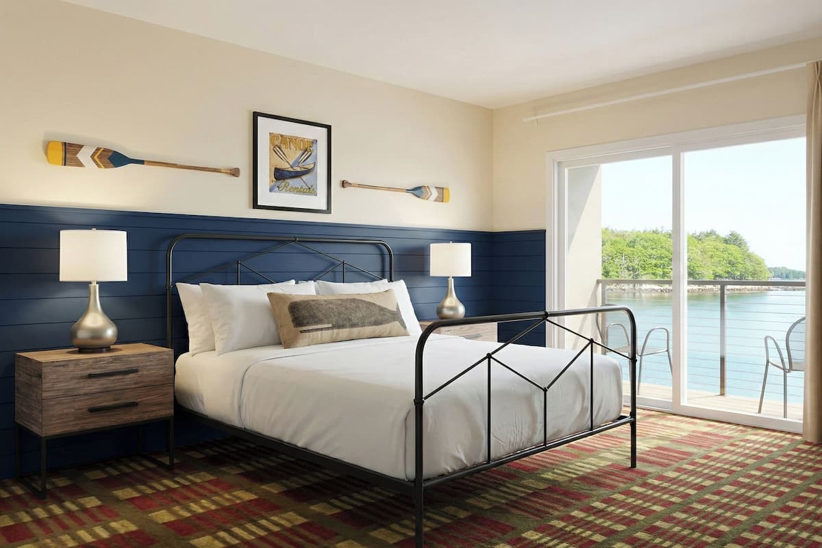 A bedroom with a bed, two bedside tables, a window with a view of a body of water, and a framed picture on the wall.