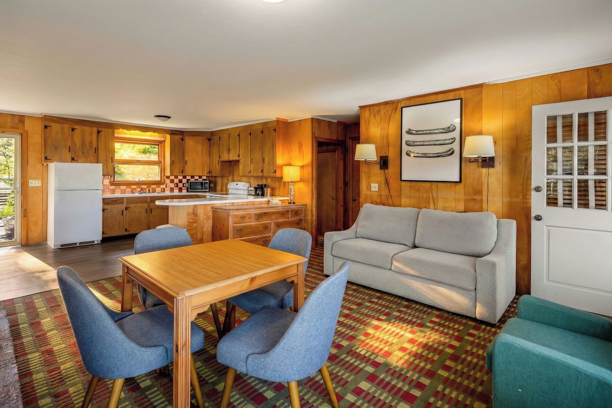 A compact kitchen with wooden cabinets, a dining table with blue chairs, and a grey sofa.