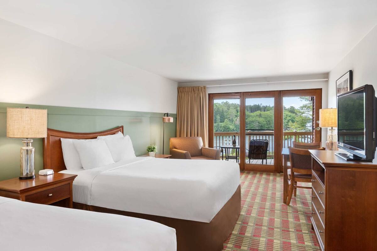 A hotel room with two beds, a desk with a TV, a balcony with a view of the forest, and a patterned carpet.