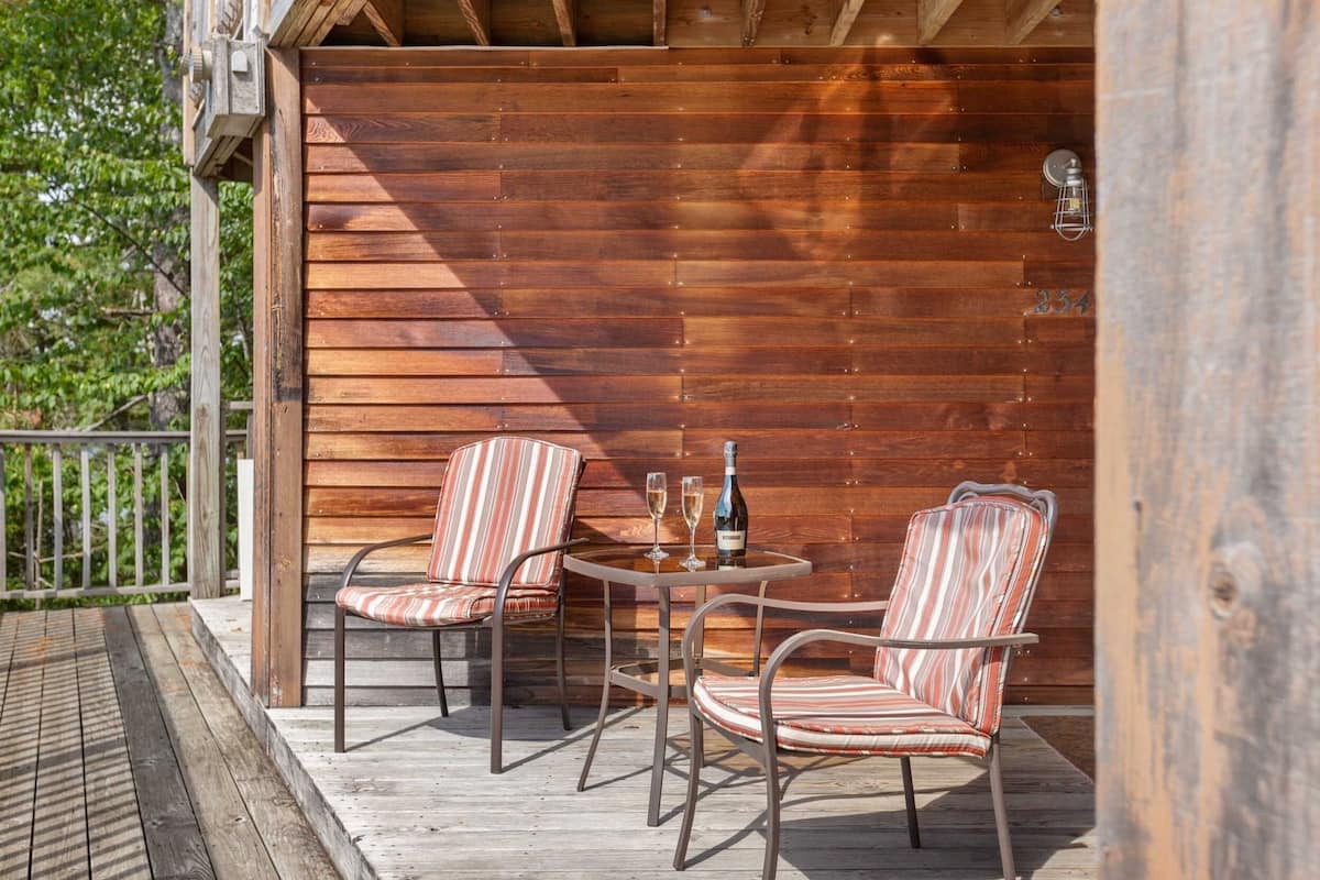 A wooden deck with two striped chairs and a table, a bottle of wine, and a lantern on the wall.