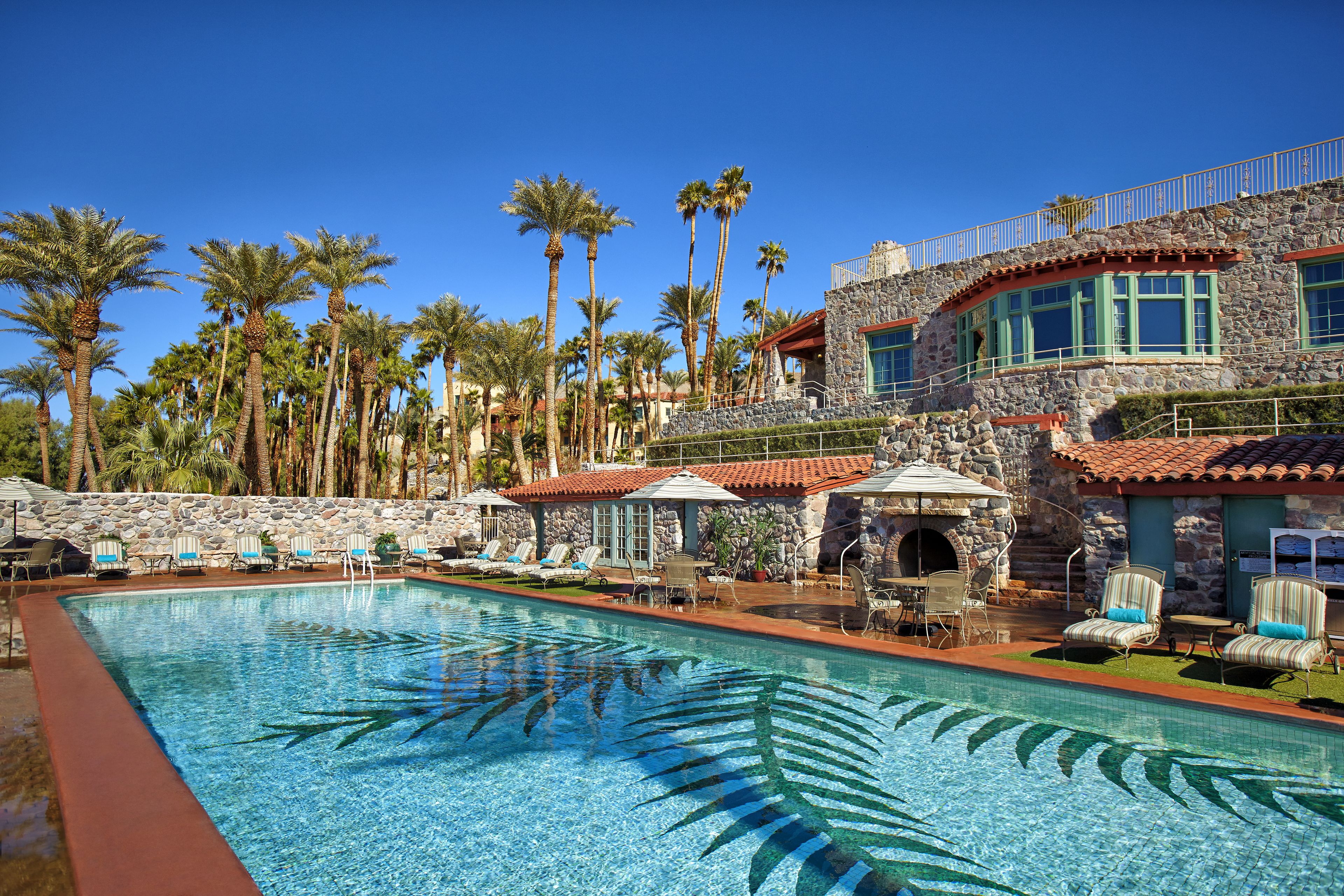 A pool area with lounge chairs, a stone building with a red-tiled roof, and palm trees.