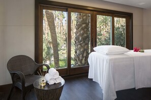 A hotel room with a bed, a chair, and a small table with towels and a bottle.