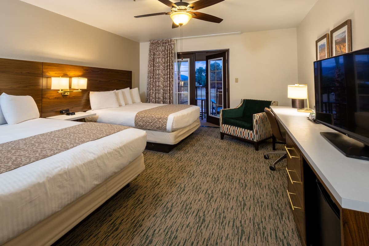 A hotel room with two beds, a desk with a TV, a chair, and a view of the outdoors through a door.