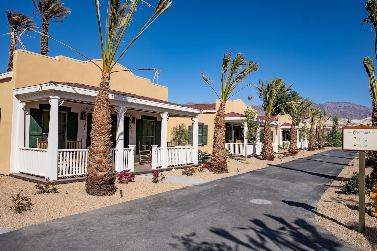 A row of single-story buildings with a white facade and a covered porch, surrounded by palm trees and a clear blue sky.