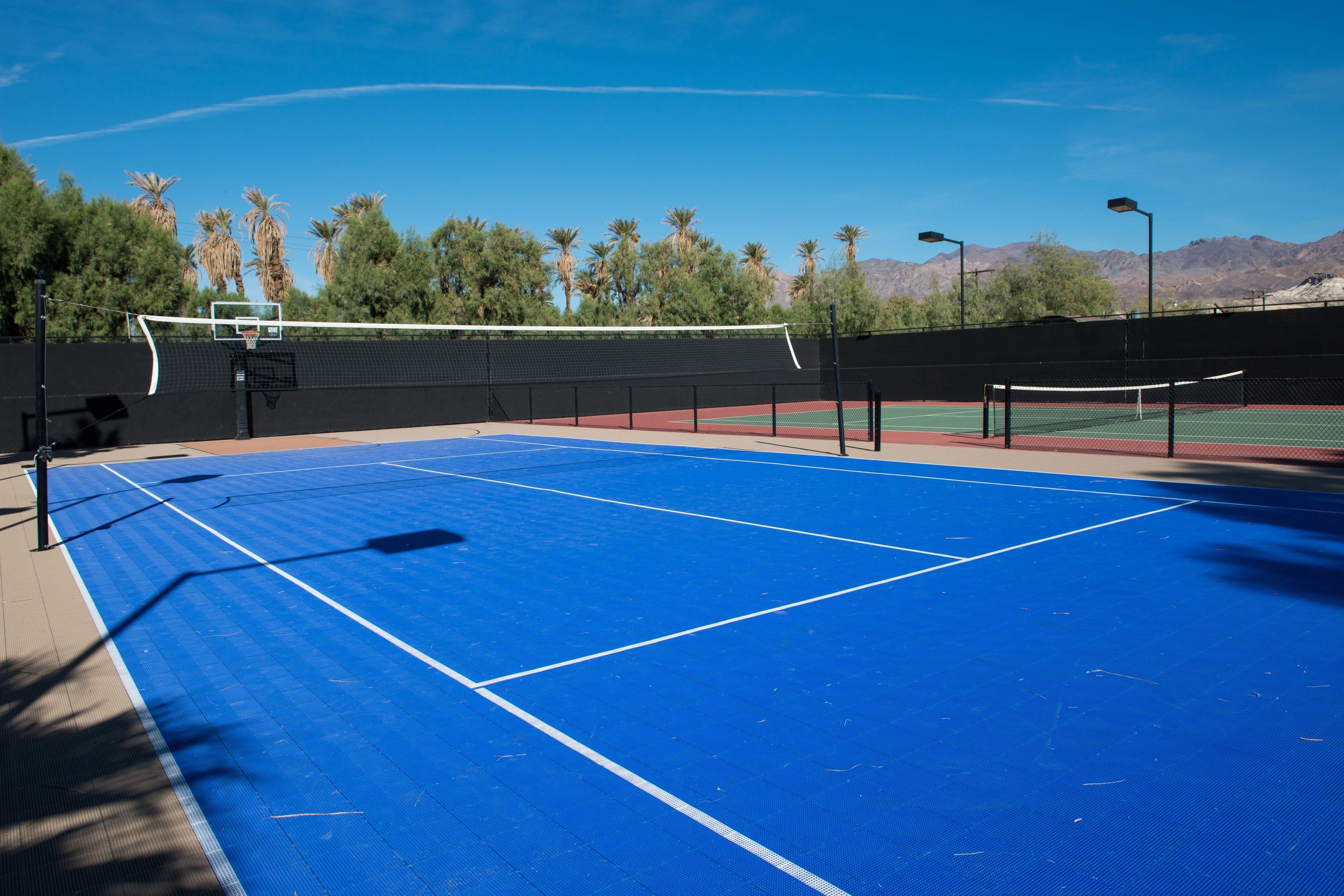 An outdoor tennis court with a blue surface, surrounded by trees and mountains in the background.