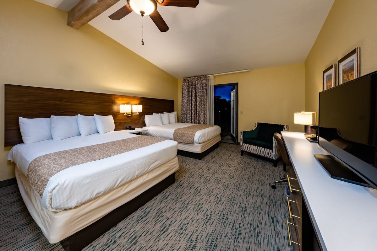 A hotel room with two beds, a desk, a chair, a television, and a ceiling fan.