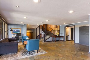 Un espacio habitable amplio con suelo de piedra, un sillón azul, un sofá, una mesa de centro y una escalera que conduce a otro nivel.