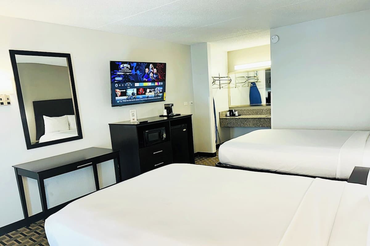 A hotel room with two beds, a TV mounted on the wall, a desk with a mirror, and a small kitchenette.