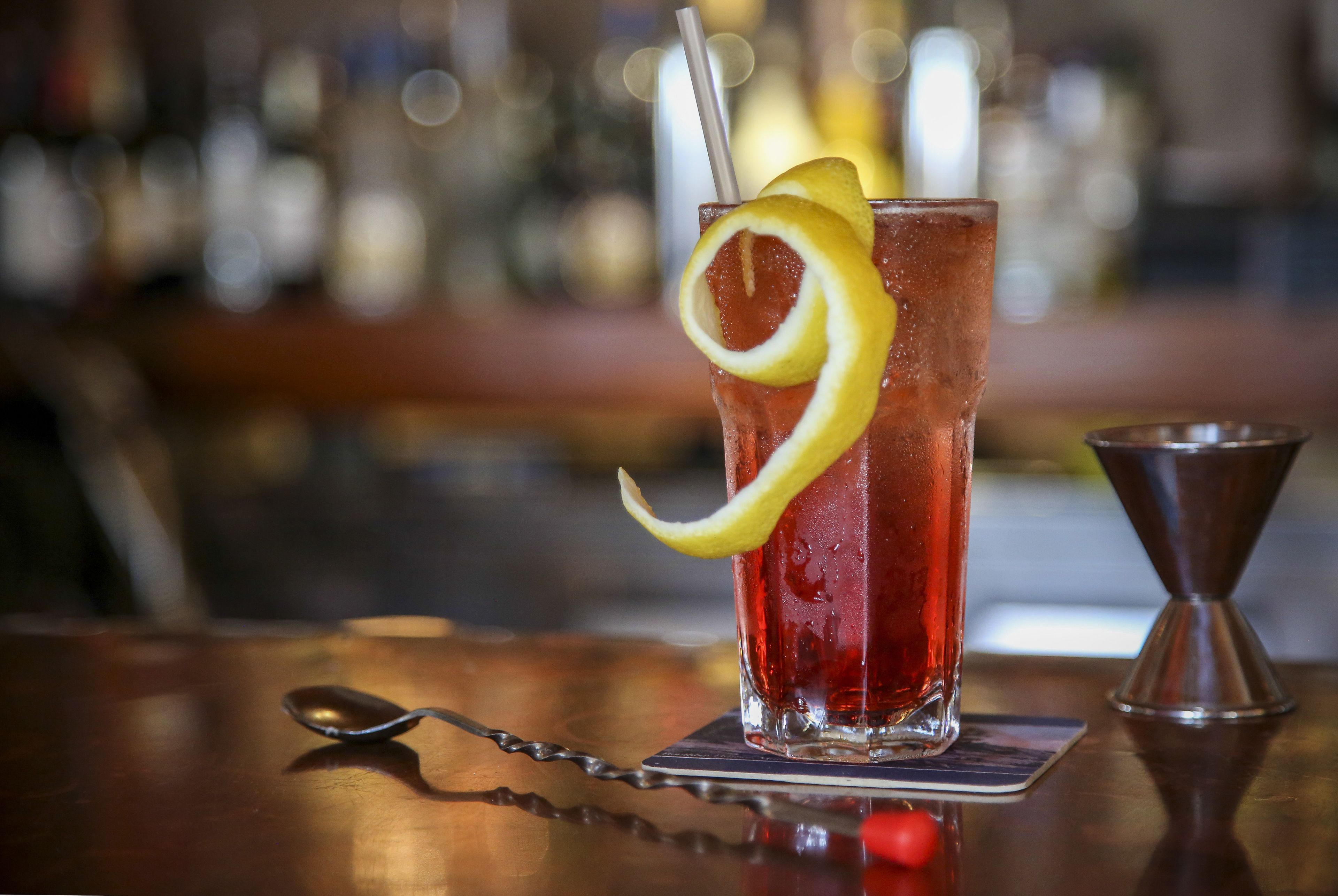 A glass of red beverage with a lemon slice and straw, placed on a coaster with a spoon beside it.
