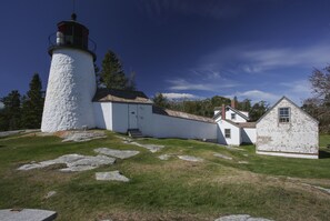A lighthouse with a white exterior and a black top, adjacent to a white building with a red roof, set against a clear blue sky.