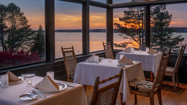 A dining area with tables set for two, large windows offering a view of a sunset over a body of water, and wooden chairs with cushioned seats. - Spruce Point Inn (Boothbay Harbor)