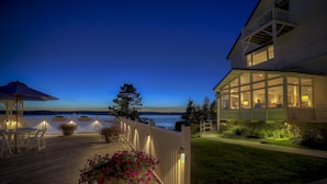 A wooden deck with outdoor seating and potted plants, overlooking a body of water at dusk. - Spruce Point Inn (Boothbay Harbor)