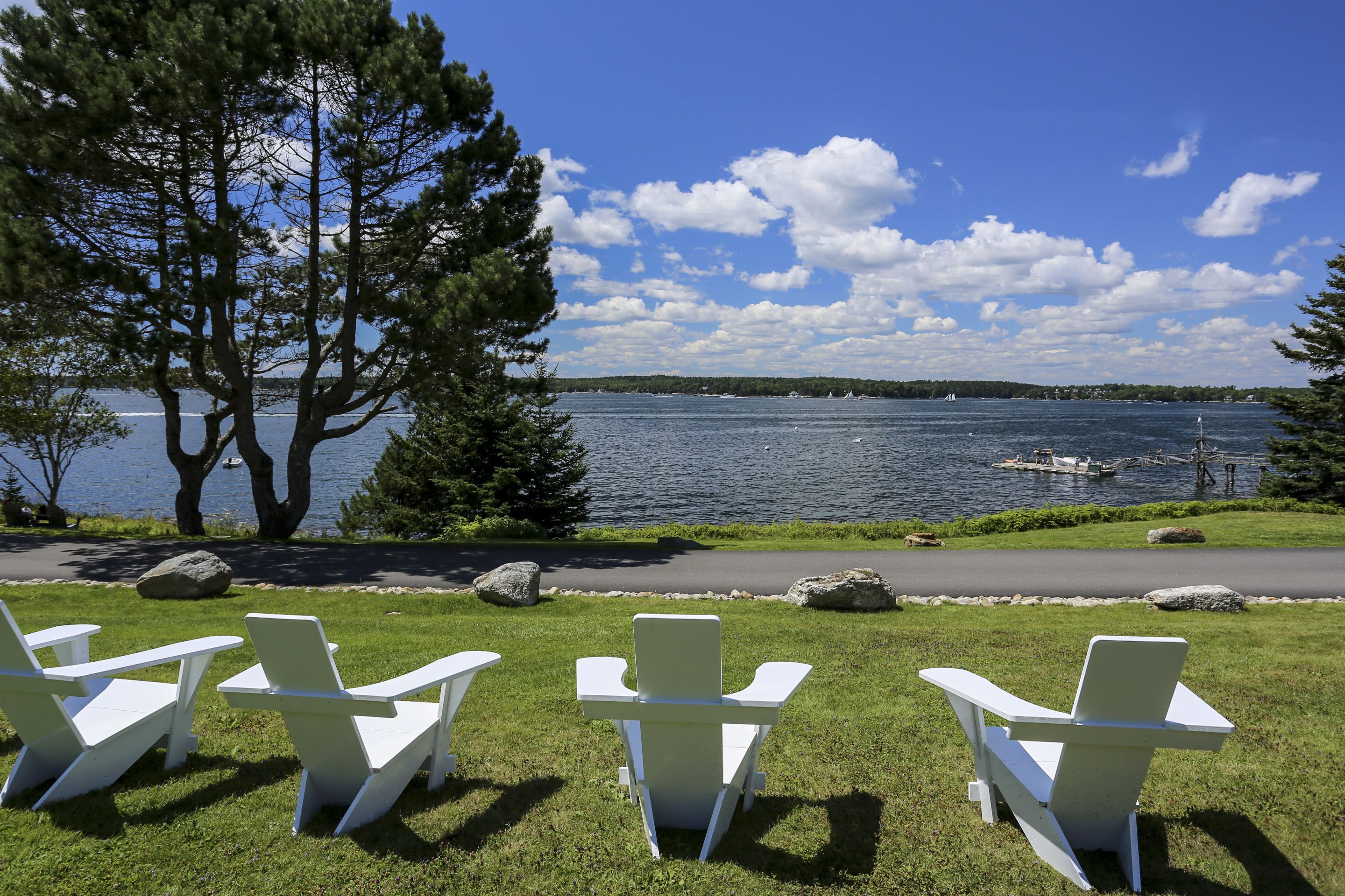 A lakeside view with three Adirondack chairs facing the water, a paved path, and a dock with boats.