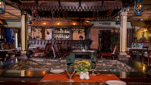 Restaurante com área de bar, cadeiras de madeira, mesa para dois com um vaso de flores e teto decorativo.