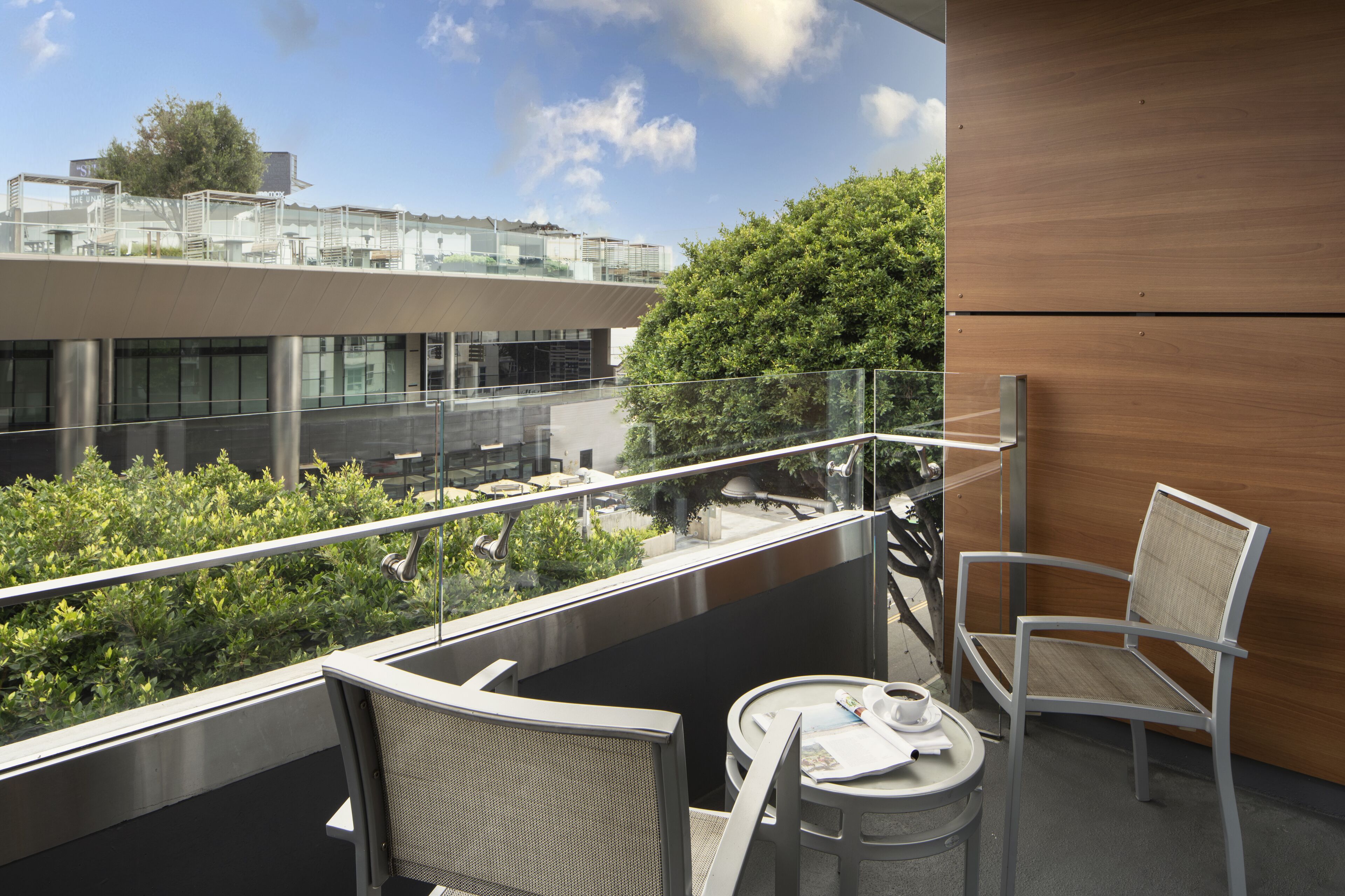 En balkon med gelænder af glas, to stole, et lille rundt bord og udsigt til en bygning og grøn vegetation.