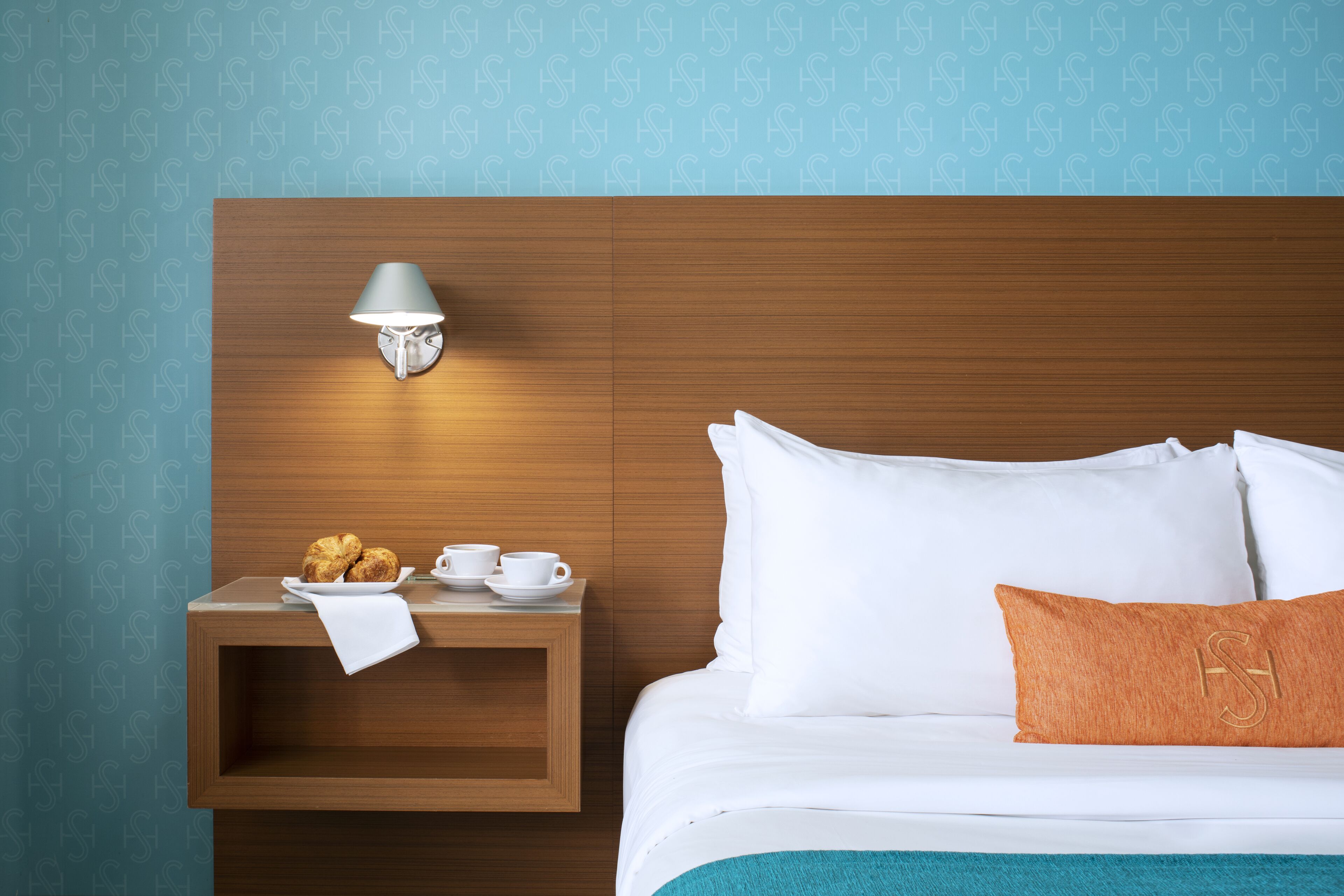 Habitación de hotel con cabecera de madera, una mesita de noche con lámpara, cama con ropa blanca y almohada naranja, y pared con un patrón repetido.