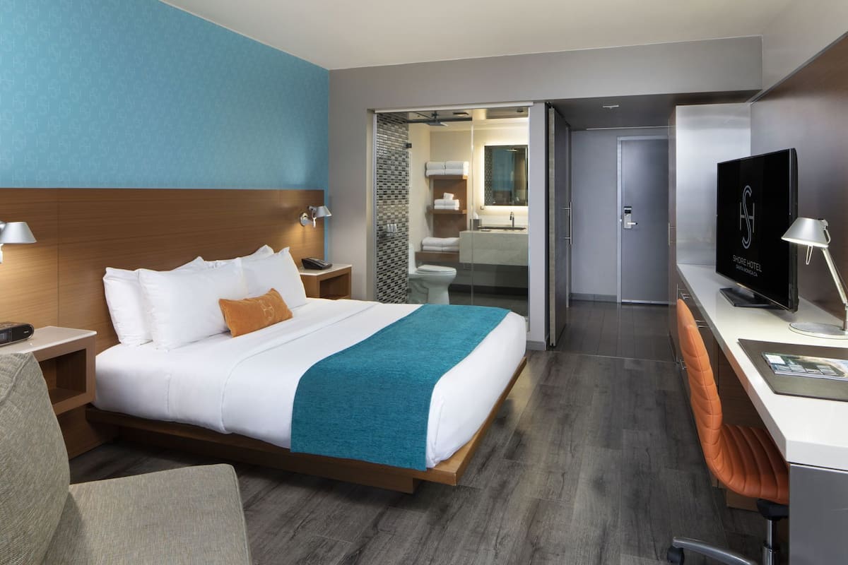 Habitación de hotel con cama, mesitas de noche, televisor, escritorio y baño visible a través de una puerta abierta.