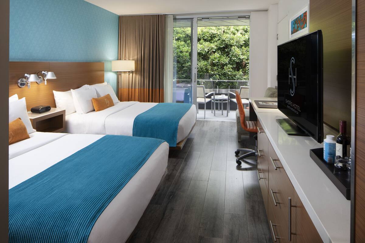 Habitación de hotel con dos camas, televisión, escritorio y balcón con vista a zonas verdes.